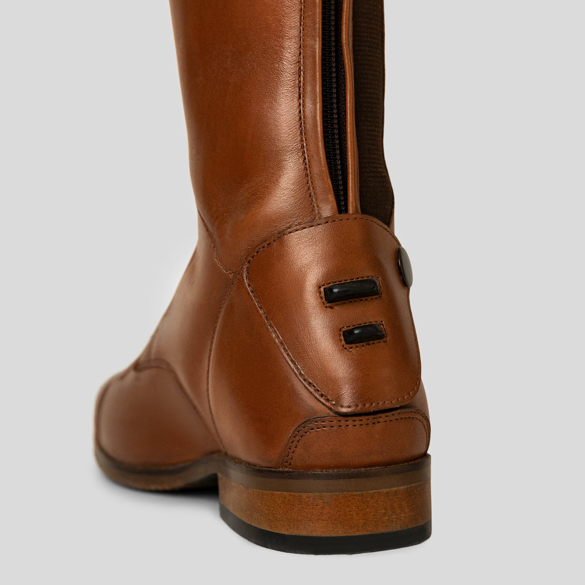 Close-up rear view of a tall brown leather boot with a back zipper and heel details, shown on a light grey background.